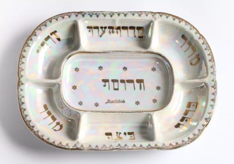 Biały talerz w kształcie owalnym. Widoczne złote napisy w języku hebrajskim w wyodrębnionych polach.