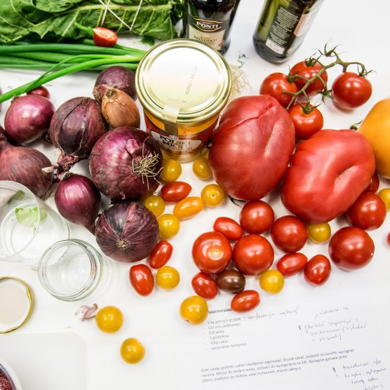 Na stole znajduje się wiele różnych warzyw z przewagą tych czerwono żółtych np. duże malinowe pomidory, mniejsze czerwone pomidory na gałązce, żółte podłużne pomidory oraz czerwone cebule